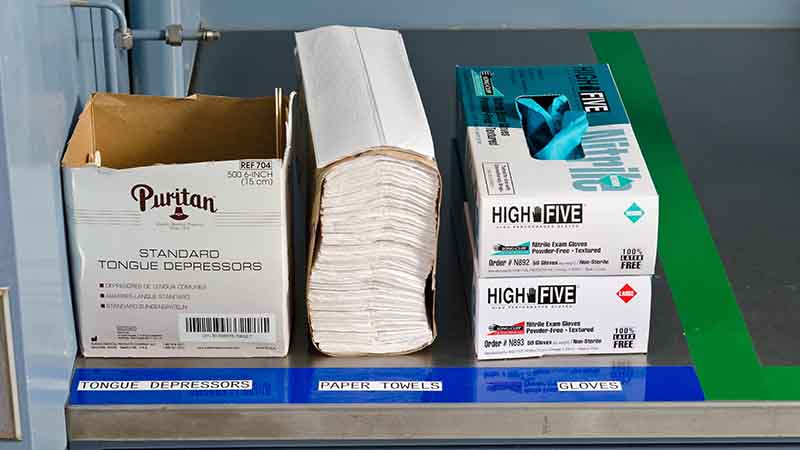 An organized laboratory table arranged according to lean 5S principles, featuring neatly placed tongue depressors, napkins, and gloves. Each item is clearly labeled, facilitating easy identification and access.